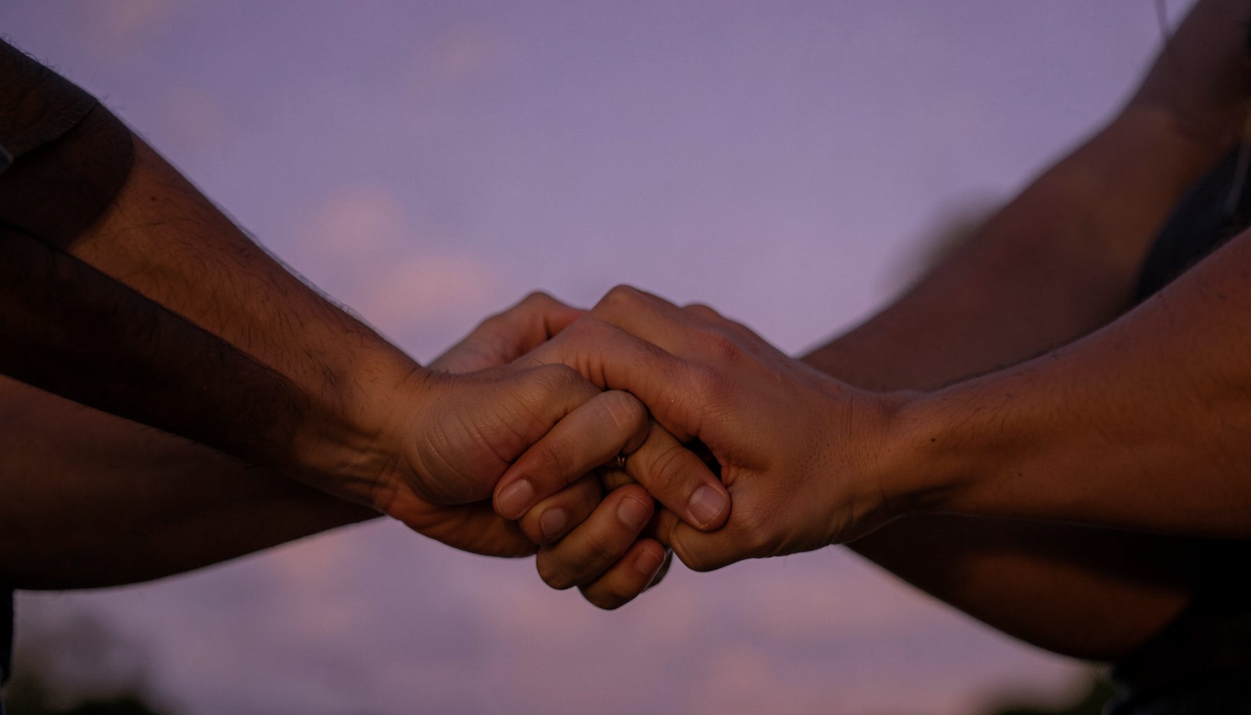 A warm, softly lit scene of hands joined in a circle against a dusky lavender sky, conveying community, hope, and quiet determination.